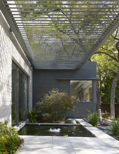 Modern house exterior with a pergola and large windows overlooking a small pond with plants. Trees are visible in the background.
