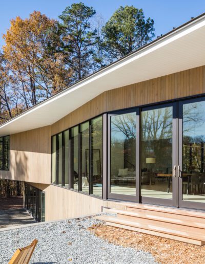 Modern house with large glass windows, sloped roof, and wooden siding, surrounded by trees. Outdoor stairs lead from gravel area to entrance.