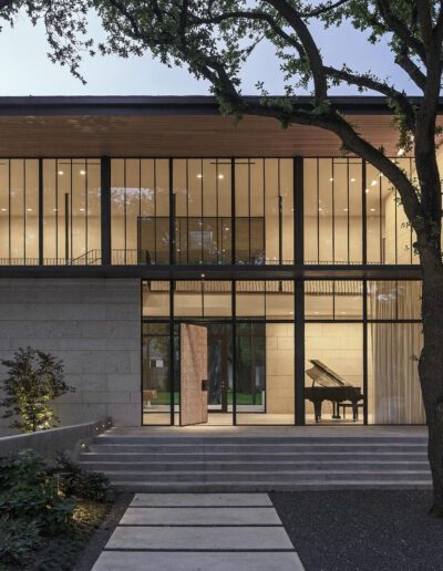 Modern house illuminated at twilight with large windows, a grand piano visible inside, and surrounded by trees.