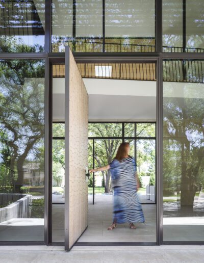 Woman walking through a modern glass door into a sunlit building with reflections of trees.
