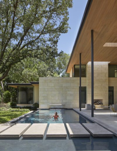 Child playing in a modern backyard pool with stone walkway and surrounding lush greenery.