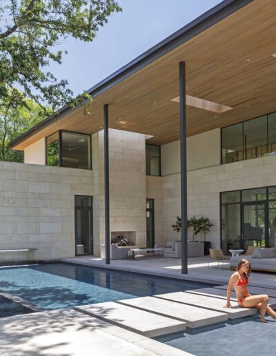 A modern house with large glass windows and a swimming pool, with a person sitting by the poolside.