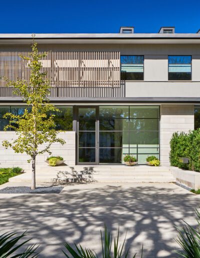 Modern two-story house with a flat roof, large windows, and a wooden accent facade. A tree and shrubs are in the front yard, and there is a paved walkway leading to the entrance.