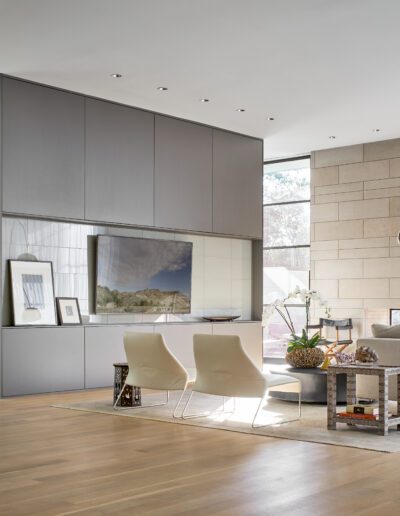 Modern living room with gray cabinets, two white chairs, a beige sectional sofa, and a floor lamp. Wooden flooring and large windows create an open, airy atmosphere.