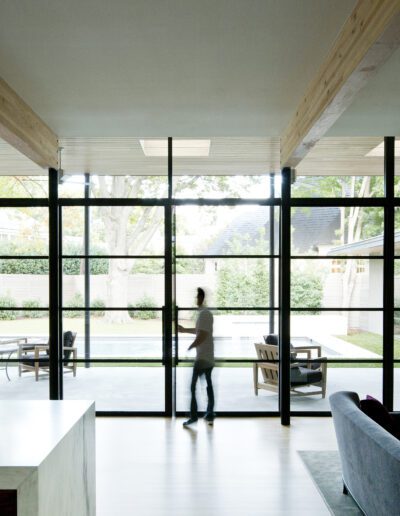 Spacious modern living room with large glass windows overlooking a patio and pool. A bowl of lemons sits on a kitchen counter. People are seen walking outside.