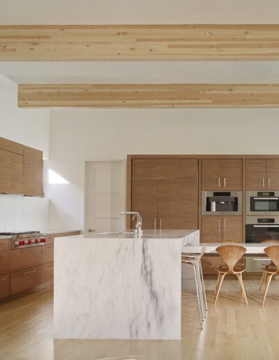 Spacious kitchen with wooden cabinetry, marble island, and built-in appliances. Light wood beams and flooring enhance the bright, minimalist design. Two chairs flank a small table.