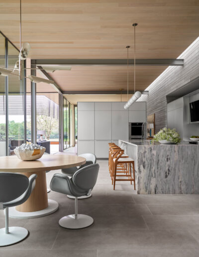 Modern kitchen and dining area with a marble island, wooden stools, round table, and swivel chairs. Large windows provide natural light, and light wood paneling covers the ceiling.