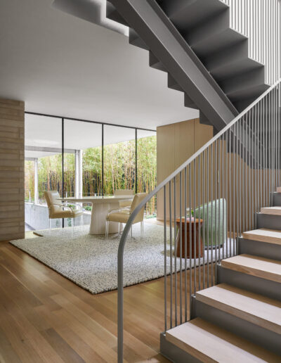 Modern interior with a wooden staircase, metal railing, and open living area. Floor-to-ceiling windows illuminate a room with a rug, chairs, and a table.