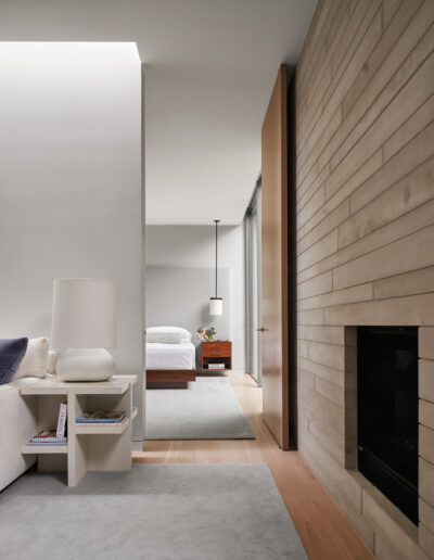 Modern living space with a partial view of a fireplace and a lamp on a side table. Through an open doorway, a bedroom with a bed, nightstand, and hanging lamp is visible.