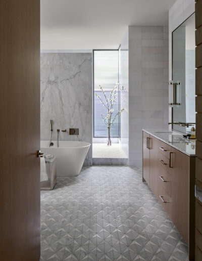 Modern bathroom with a freestanding bathtub, large wall mirror, wooden cabinets, and geometric patterned floor tiles. A floor-to-ceiling window allows natural light and features a decorative branch.